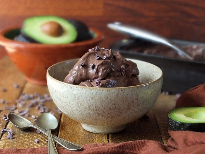 A bowl of Plant Paradox chocolate avocado ice cream sitting on a wooden table with spoons and an avocado around it