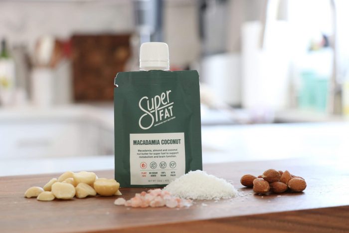 A bottle of SuperFat macadamia coconut nut butter sits on a counter with its ingredients around it.