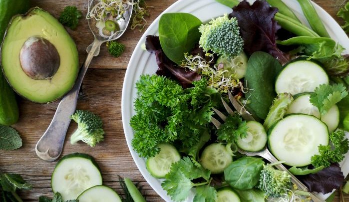 A white plate of various green vegetables sitting on a wooden surface with a spoon sitting next to it.