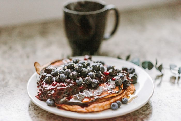 A plate of Plant Paradox blueberry pancakes next to a black coffee cup on a wooden table
